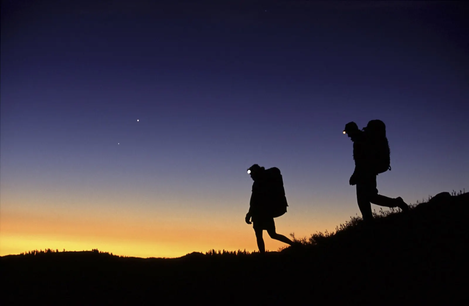 Two trekkers with headlamps setting out along a ridge at dawn